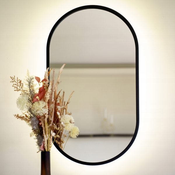 Podświetlane lustro LED w czarnej ramie, zawieszone nad konsolą z kwiatami. Lustro podświetlane LED jest dostępne w sklepie internetowym Magia Lustra – magialustra.pl.