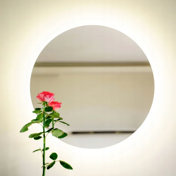 Okrągłe lustro LED bez ramy, zawieszone na jasnej ścianie z dekoracyjną rośliną. Lustro LED bez ramy jest dostępne w sklepie internetowym Magia Lustra – magialustra.pl.